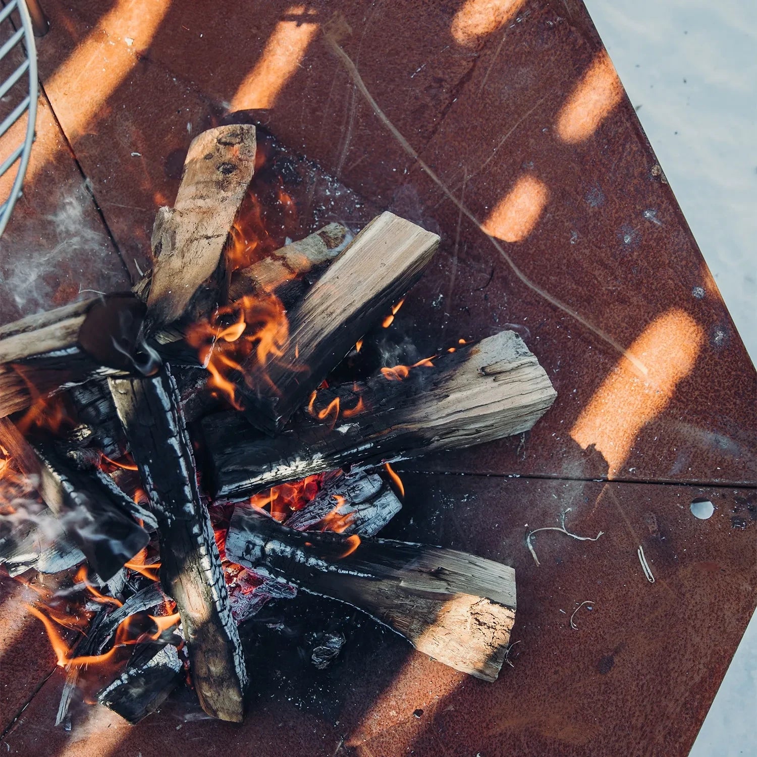Höfats TRIPLE 120 Fire Pit in Corten Steel with burning logs, placed on a white surface.